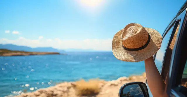 Traveler holding straw hat out car window while driving along sunlit coastline