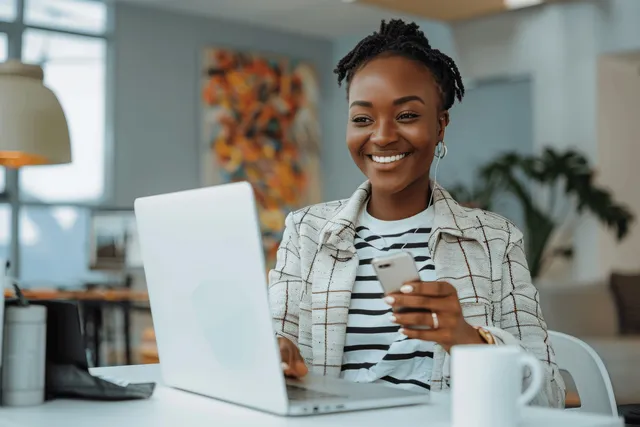 Smiling black woman using smartphone while working on laptop at office desk