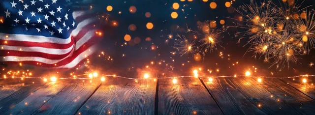 An American flag waves as sparklers and fireworks burst over rustic wooden boards for July Fourth.