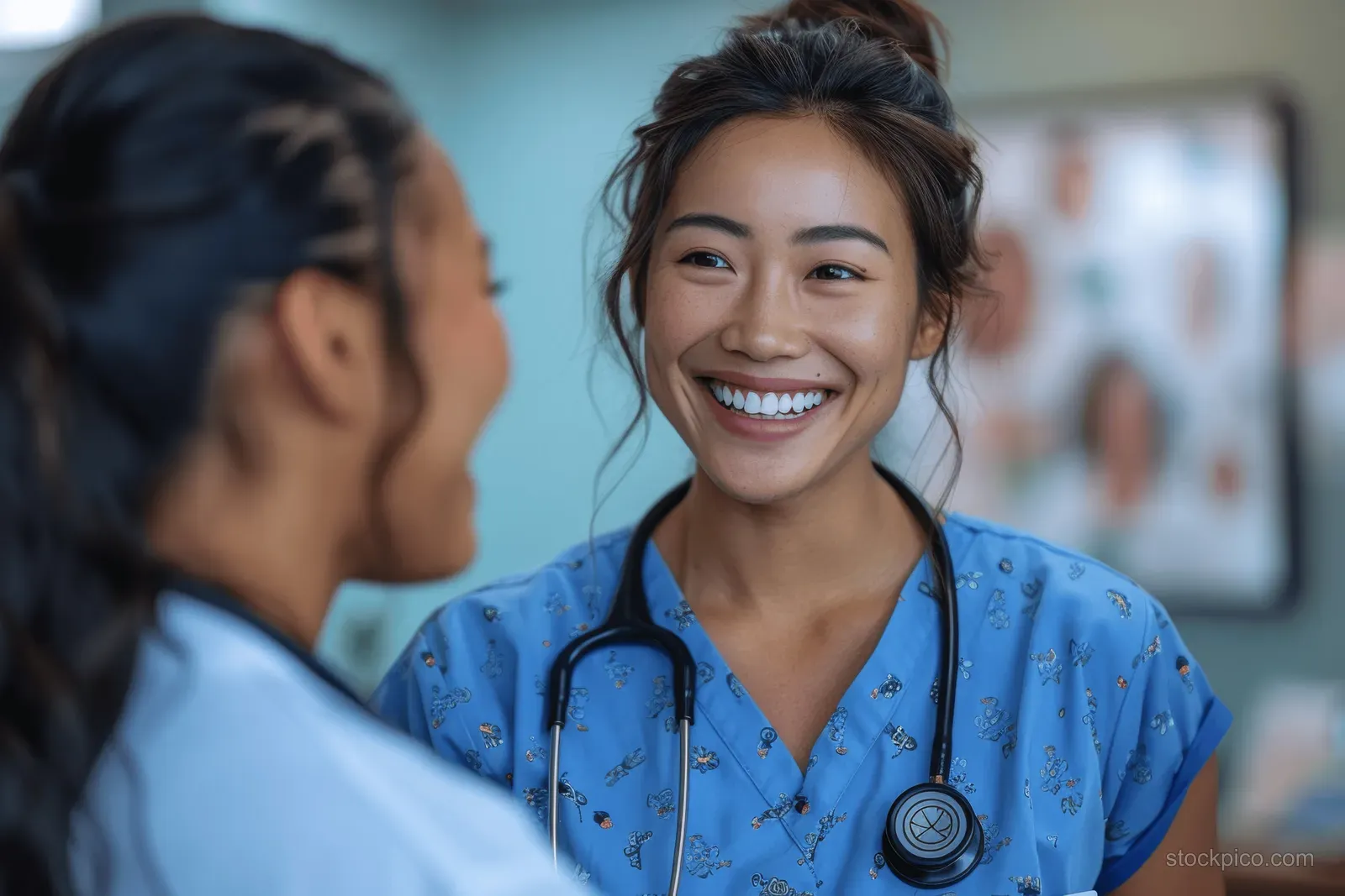 An Asian nurse smiling in blue scrubs talks with a patient in a hospital room.