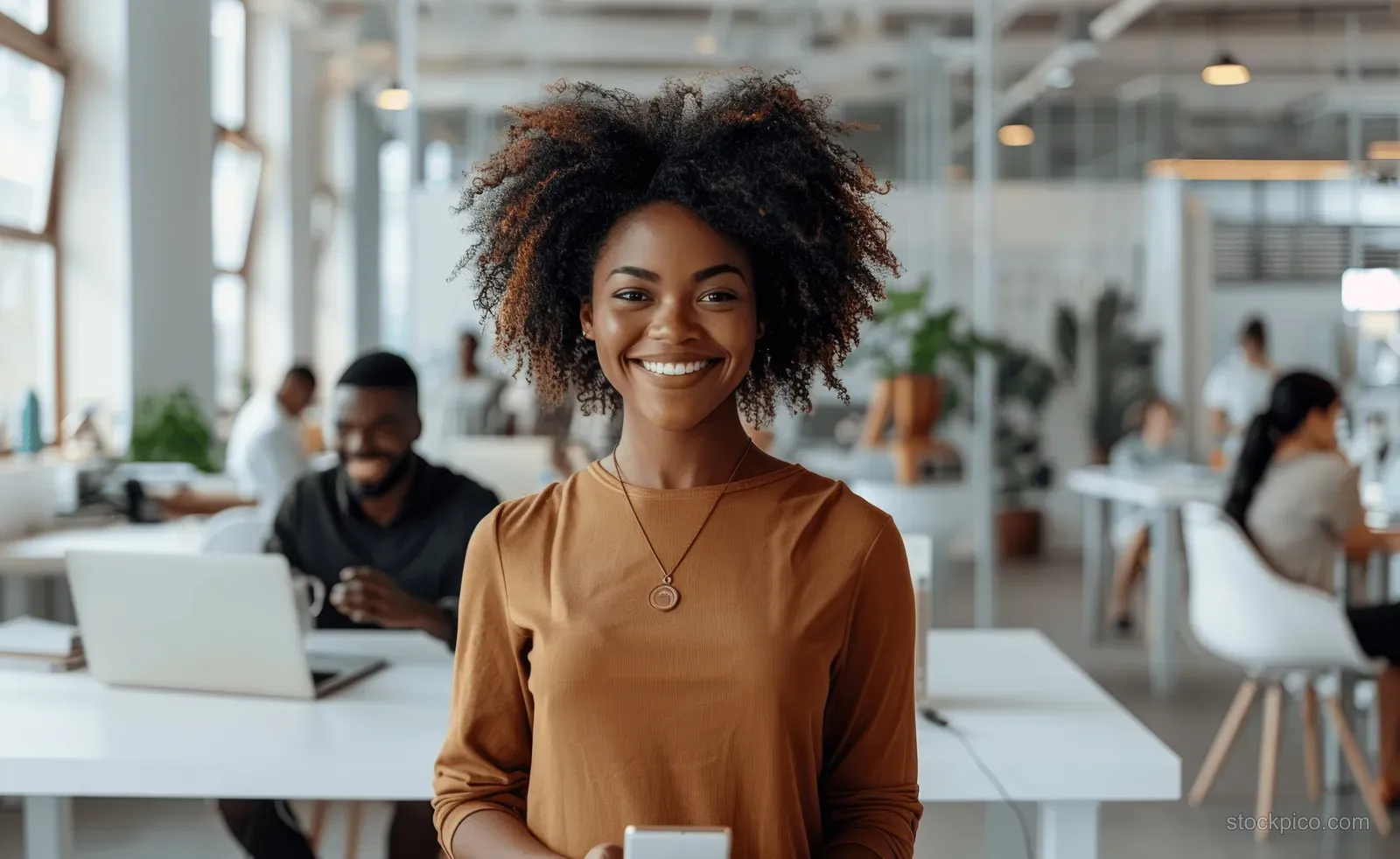 African American woman standing in a modern office while holding a smartphone and smiling in natural light.