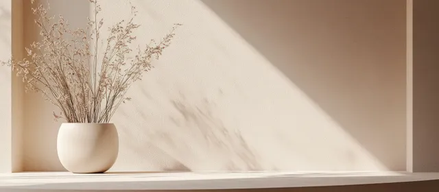 Beige vase with dried stems sitting on a recessed shelf with soft diagonal sunlight shadows.