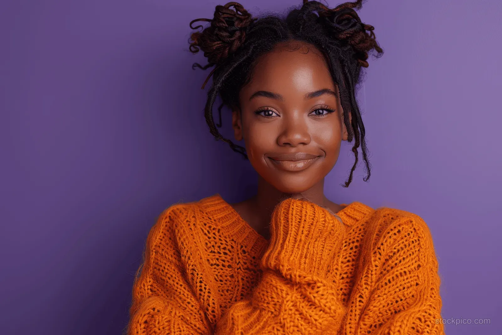 Smiling young woman in an orange knit sweater with natural two-bun hair against a purple background.