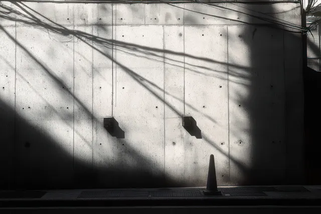 Black and white urban scene showing a concrete wall with electrical boxes, a traffic cone, and angular shadows from harsh light.