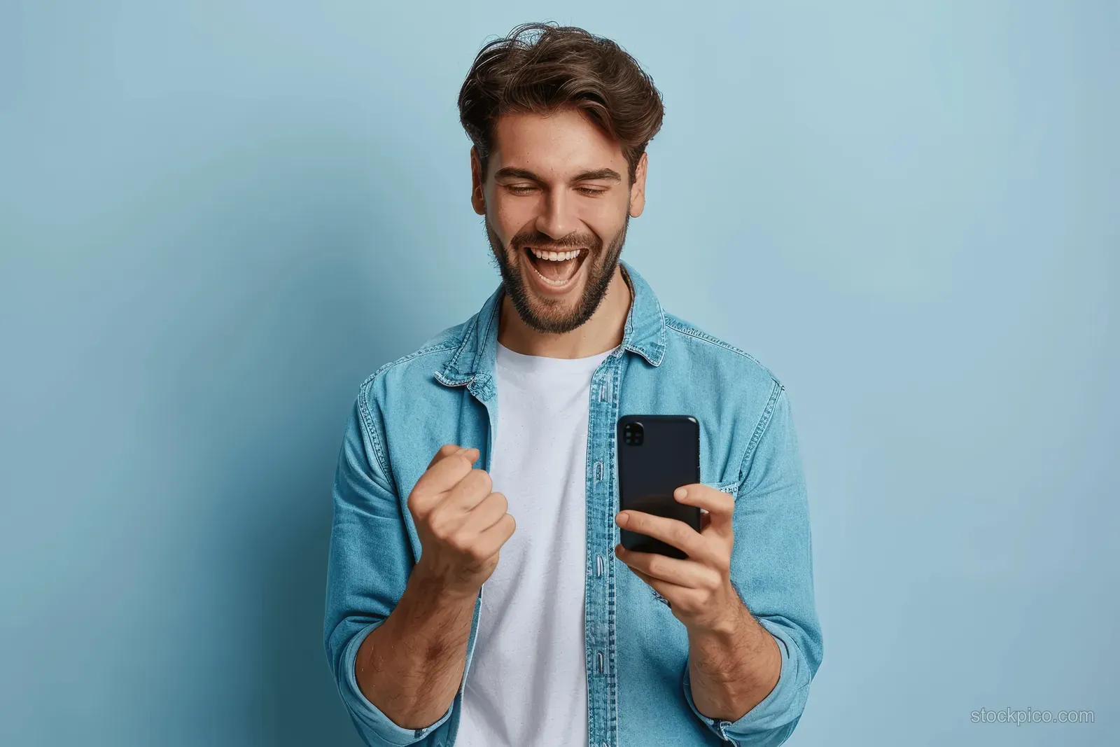 A smiling man holds a phone and celebrates with a raised fist against a light blue background.