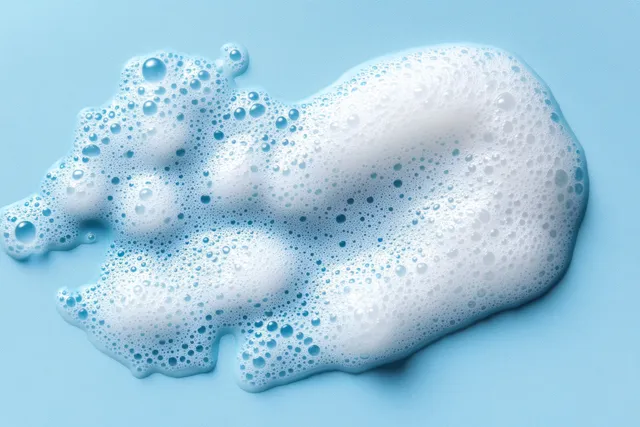 White cleansing mousse foam with small bubbles is spread across a blue background.