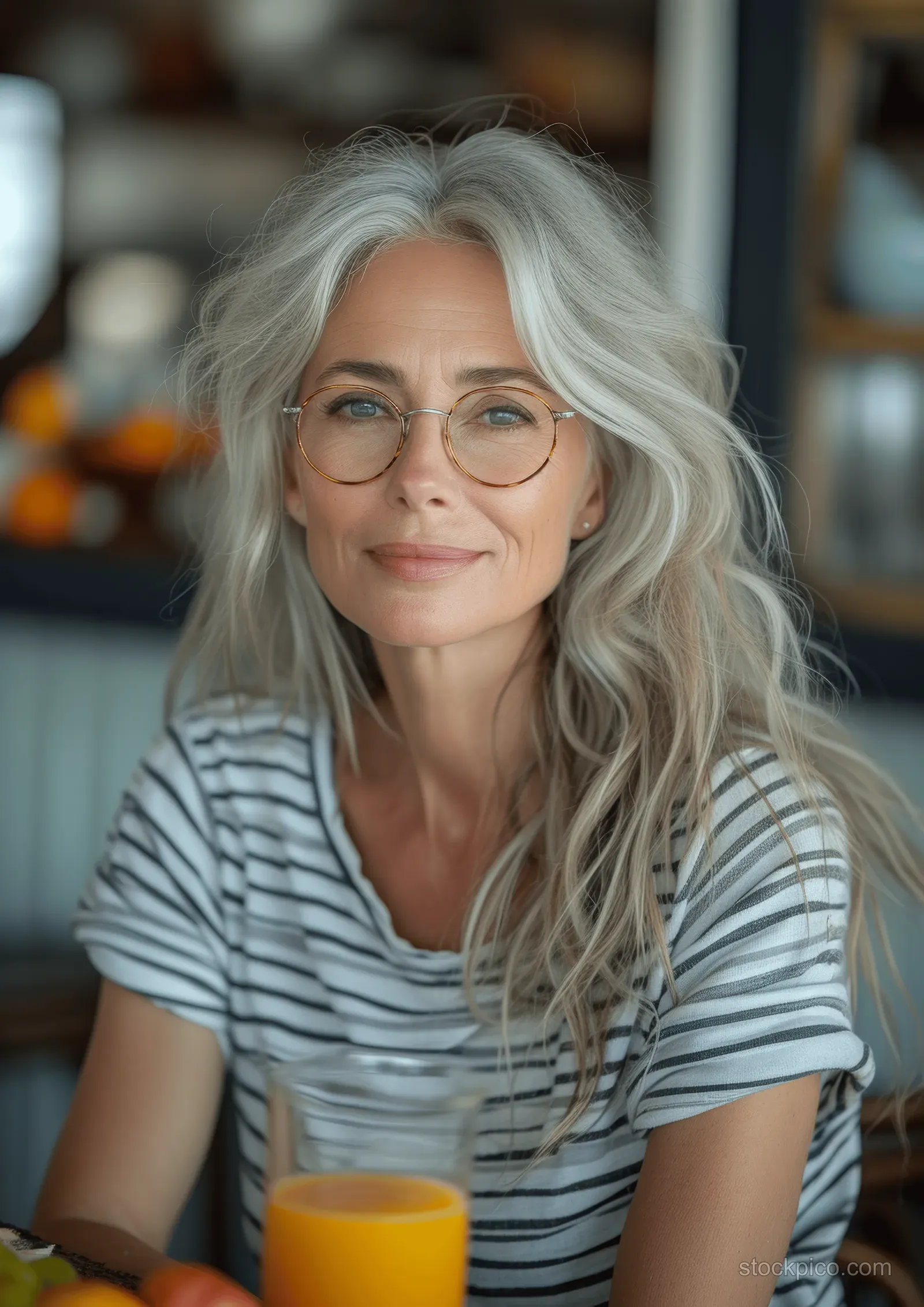 Smiling elderly woman with silver hair and glasses sits in a bright kitchen holding a glass of orange juice.