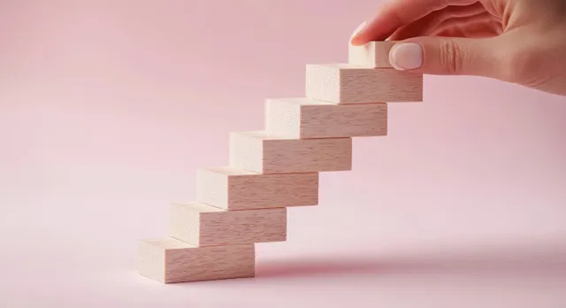Hand placing wooden blocks into an ascending staircase on a soft pink backdrop