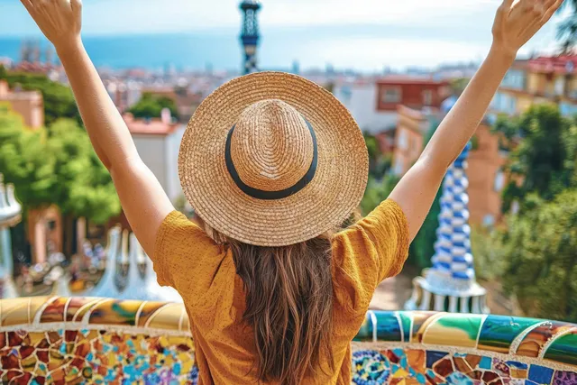 A smiling traveler raises arms for a selfie on a colorful mosaic terrace at Park Güell with Barcelona city views.