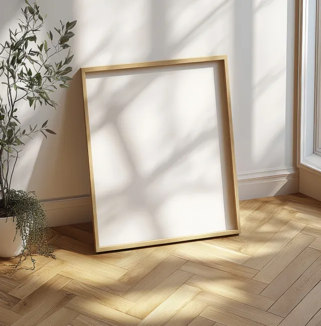 A blank picture frame leans in a minimalist Scandinavian room corner on a herringbone wood floor with soft sunlight shadows.
