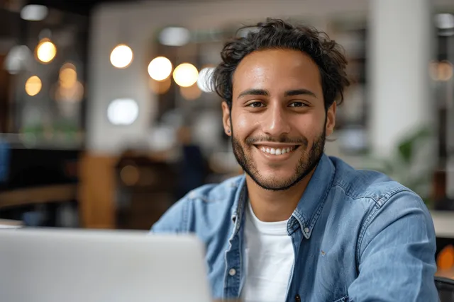 Smiling Arabian man sits at an office desk working on a laptop and looking toward the camera.