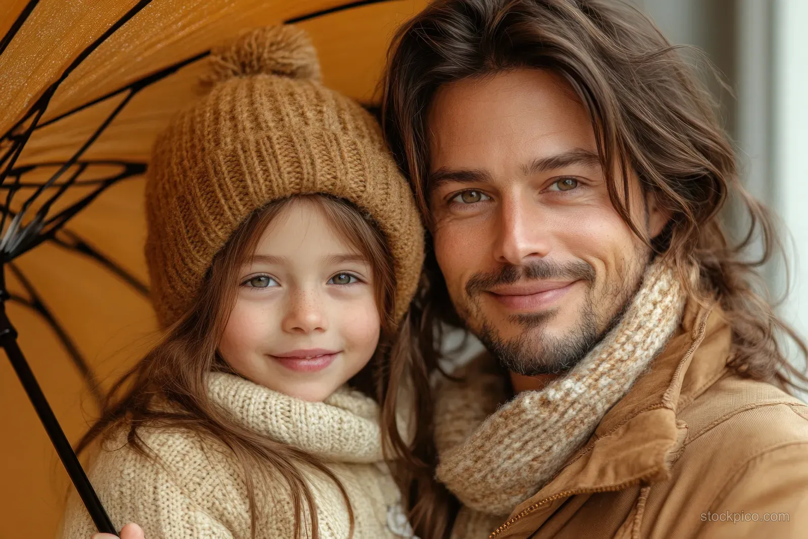 A father and daughter smile under a colorful umbrella while wearing warm knit hats and scarves on a beige background.