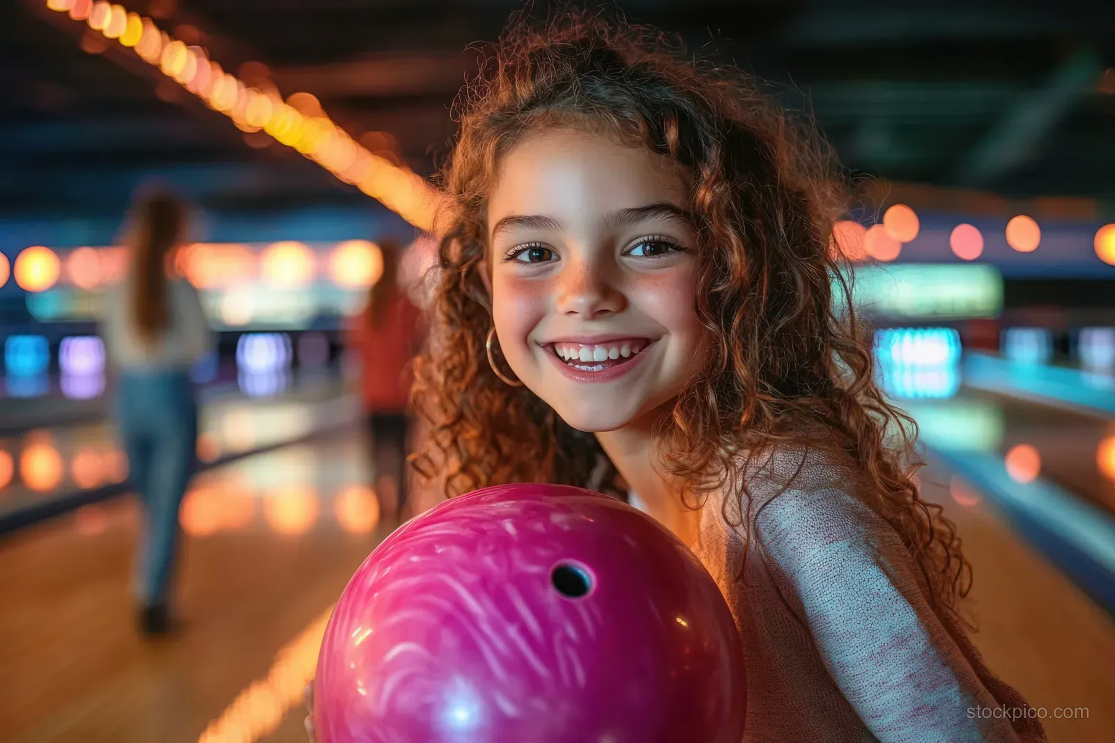 A smiling young girl holds a bright pink bowling ball in an indoor bowling alley with blurred colorful lights.