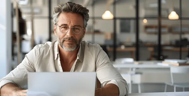 A middle-aged man with glasses researches on a laptop at a desk in a modern office with blurred glass reflections.
