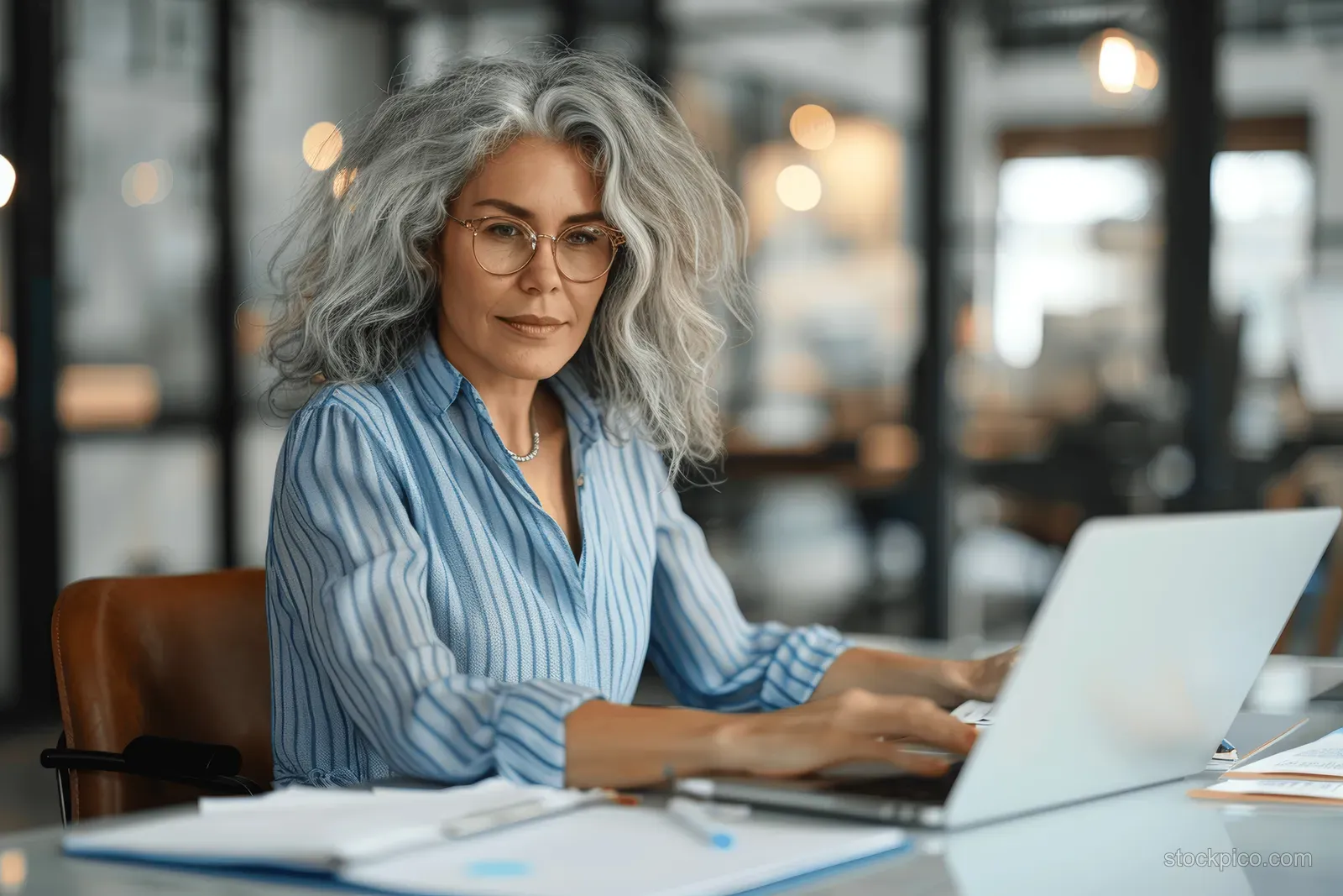 A focused woman with grey curly hair and glasses types on a laptop at a desk inside a modern glass-walled office.