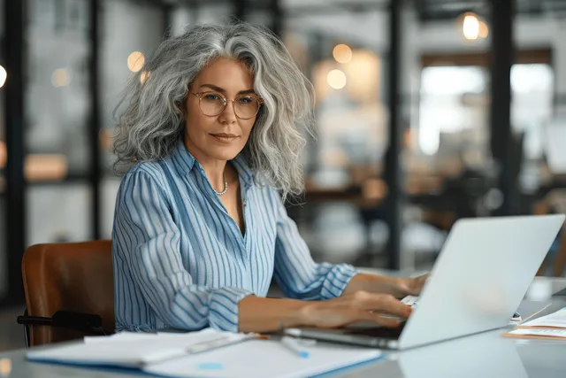 A focused woman with grey curly hair and glasses types on a laptop at a desk inside a modern glass-walled office.