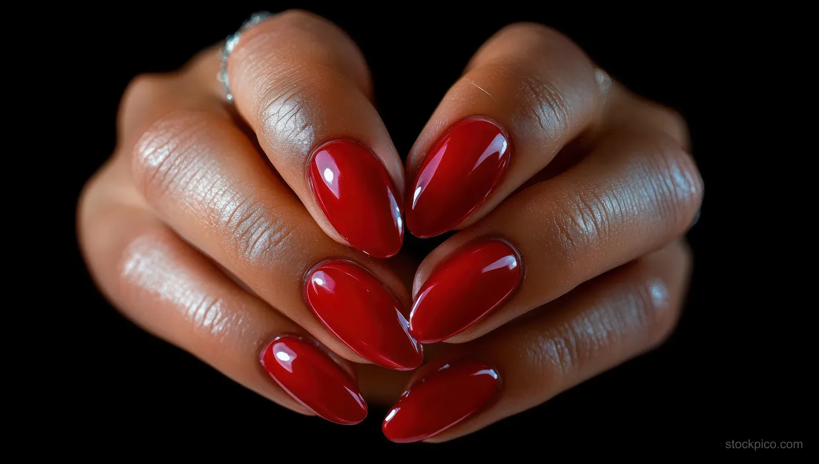 Hands with glossy red almond-shaped manicure nails and delicate rings holding fingers together on a black background.