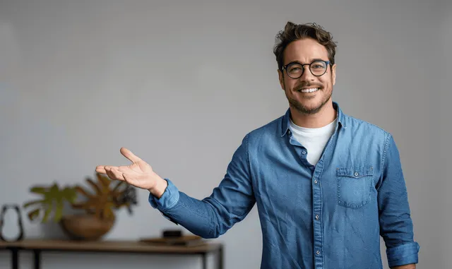A smiling man in a blue shirt extends his open palm gesture in a grey studio scene.