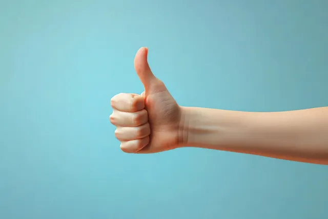 Female hand gives a thumbs up gesture against a soft pastel blue background.