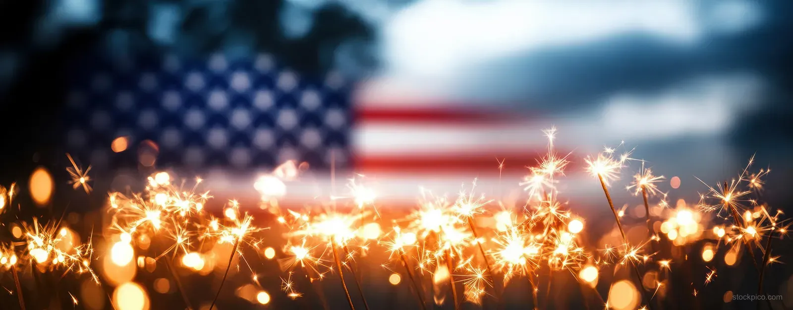 Sparklers glow in front of a softly blurred American flag with warm bokeh lights at night.