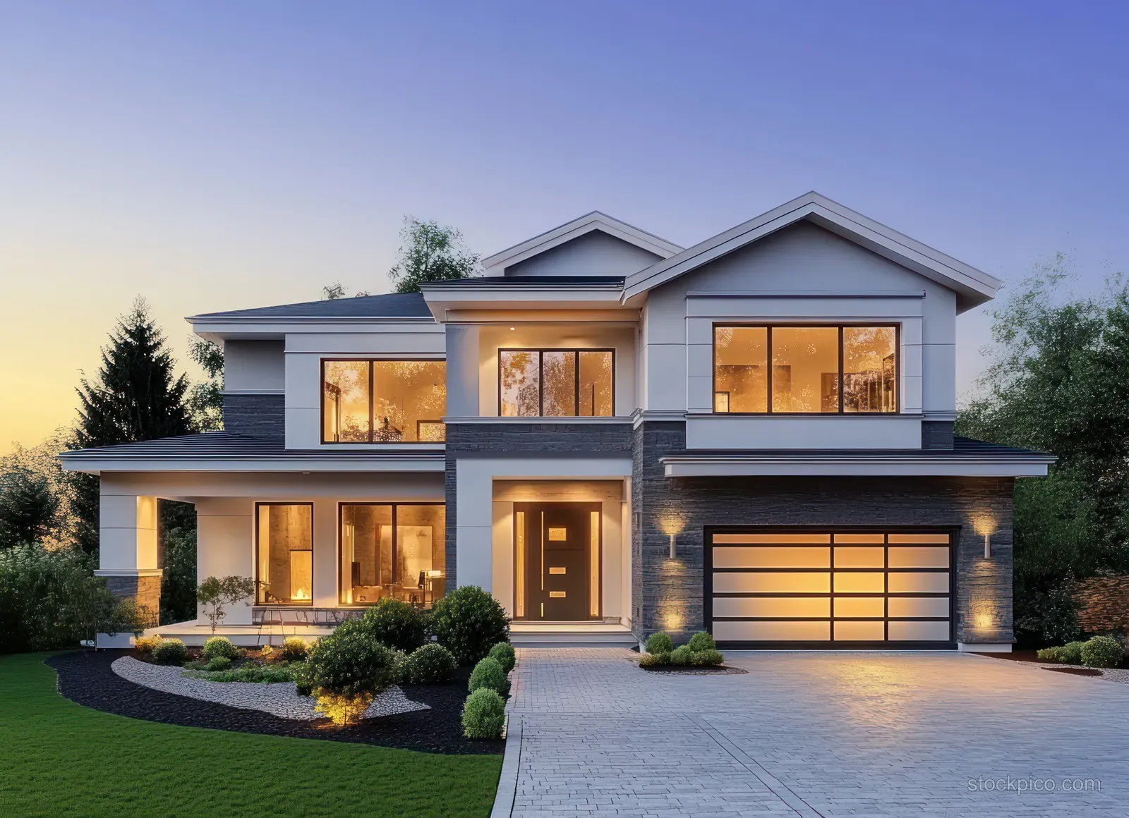 Modern house exterior at twilight with glowing large windows, lit entrance, and driveway toward the garage.