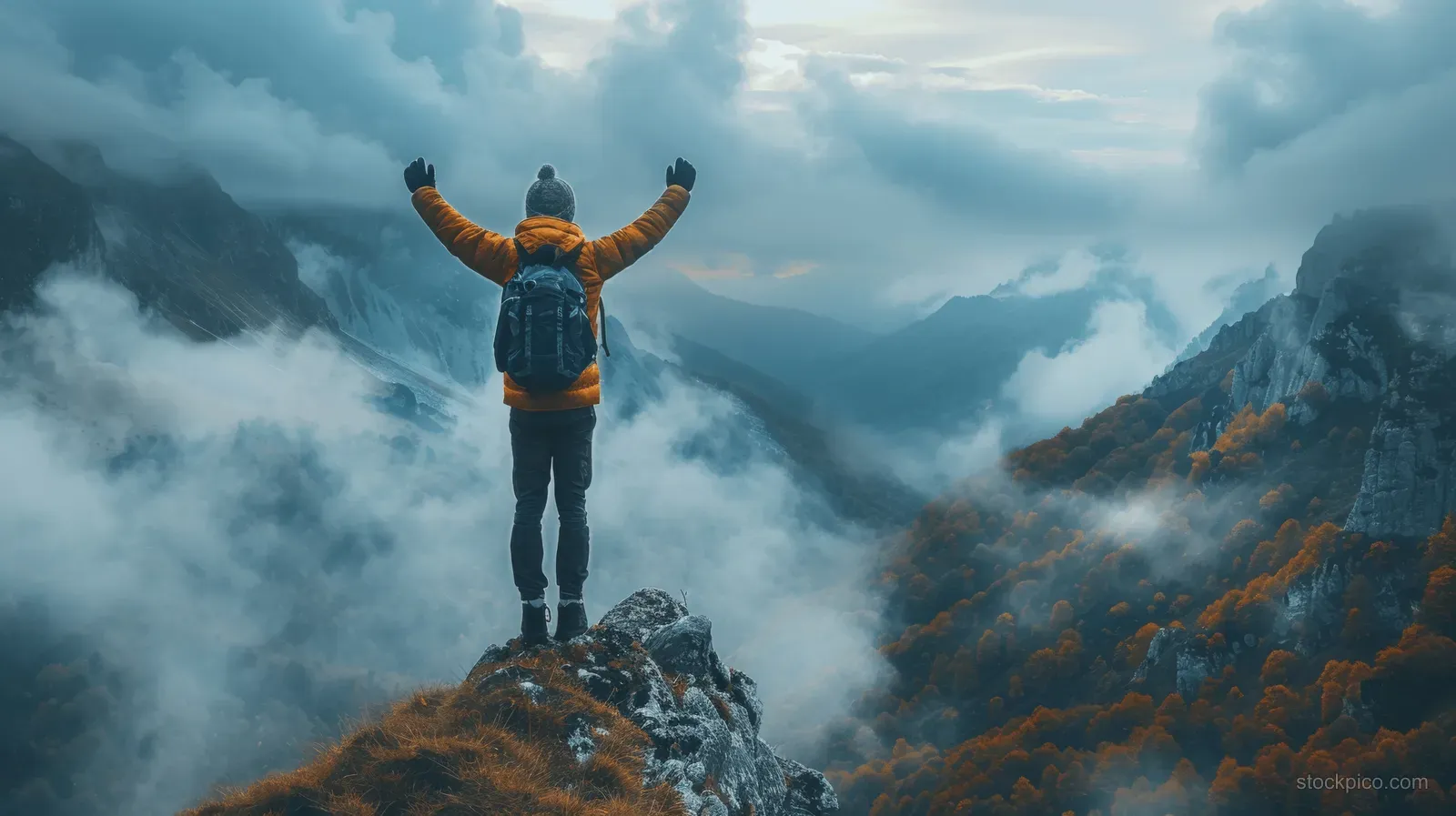 A triumphant hiker stands on a rocky mountain peak with arms raised above swirling clouds at sunrise.