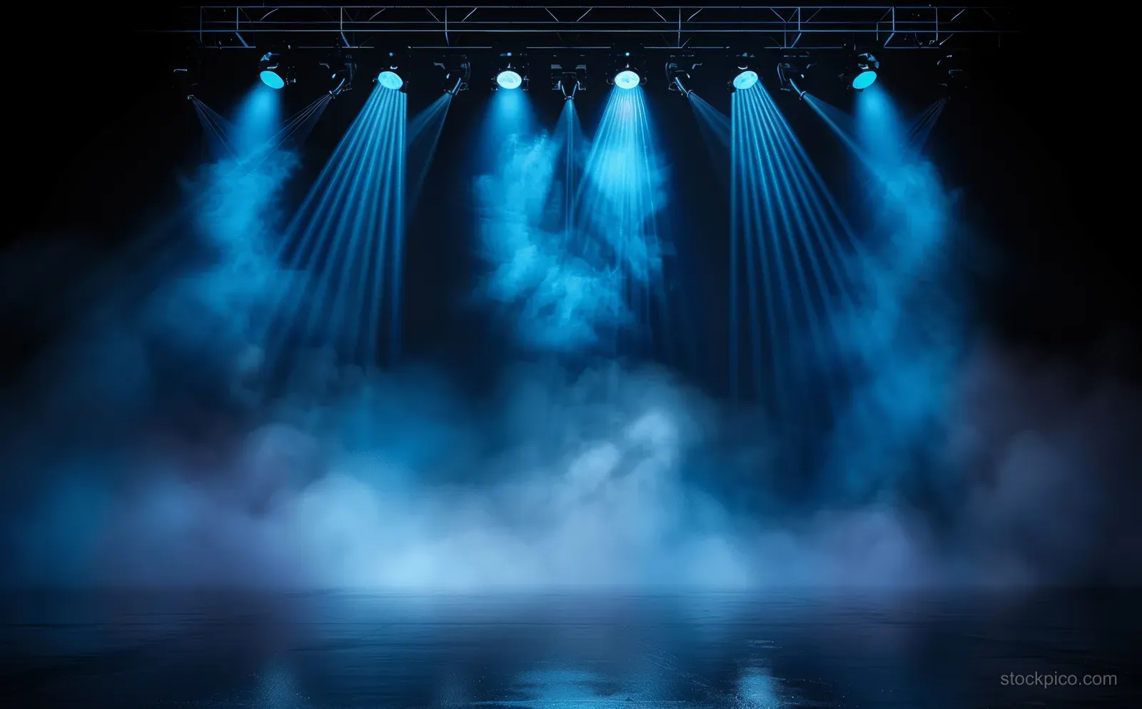 Blue spotlights shine over a concert stage with dense smoke across a dark night backdrop.