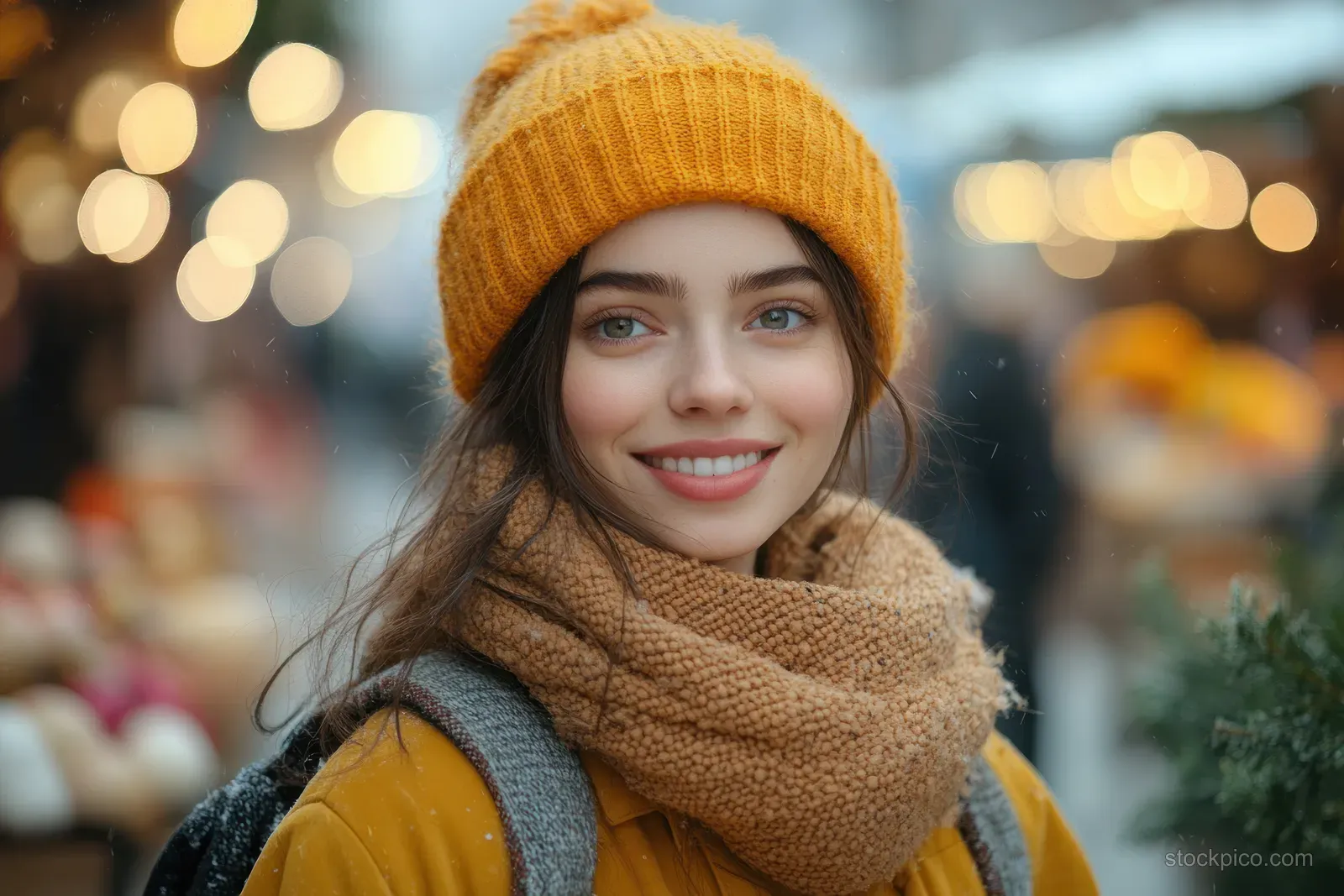 A smiling woman in a yellow coat and knit hat stands at a winter market with blurred lights behind her.