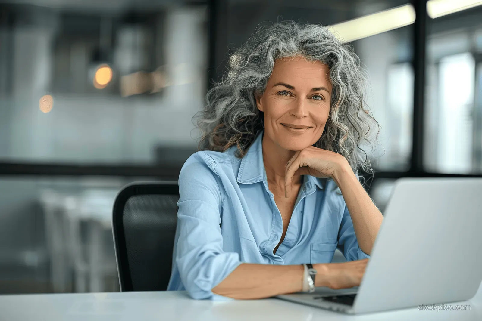 A smiling senior businesswoman works on a laptop at her desk in a modern office.