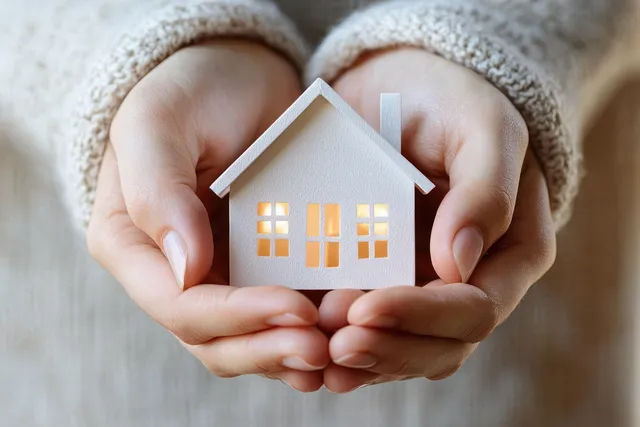A couple’s hands cradle a small white house model in warm light against a soft fabric background.
