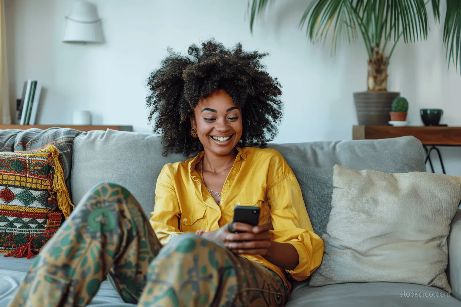 Young woman in a yellow shirt sits on a sofa at home, holding a smartphone and smiling in a bright living room.