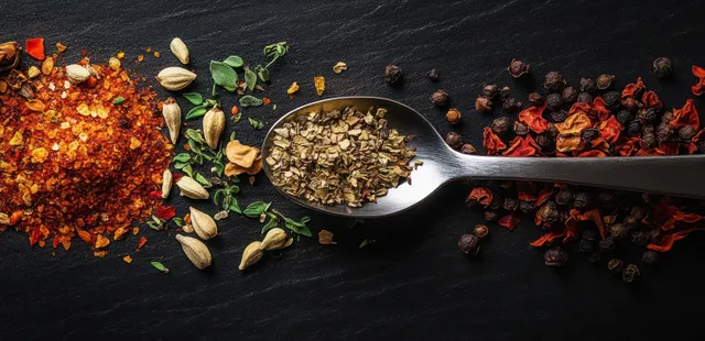 A spoon holds dried oregano as red pepper flakes, peppercorns, and other herbs scatter on dark slate.