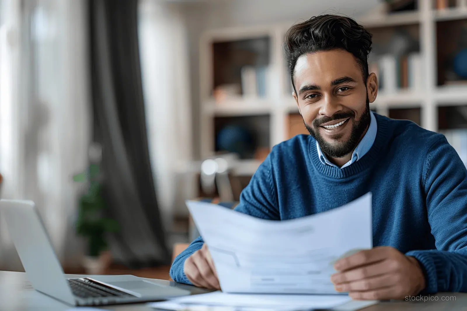 Smiling man in a blue sweater reviews documents at a desk while using a laptop in a modern home office.