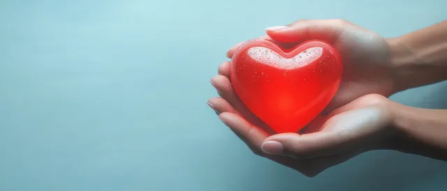 Two hands gently cradle a glowing red heart with soft light on a blue background.