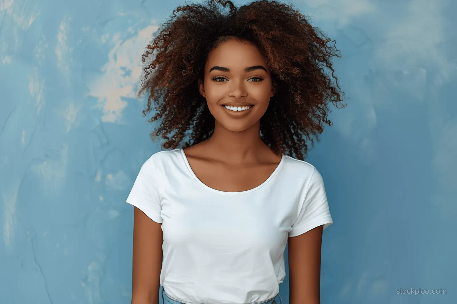 A young woman with curly hair smiles in a white T-shirt against a textured blue sky backdrop.