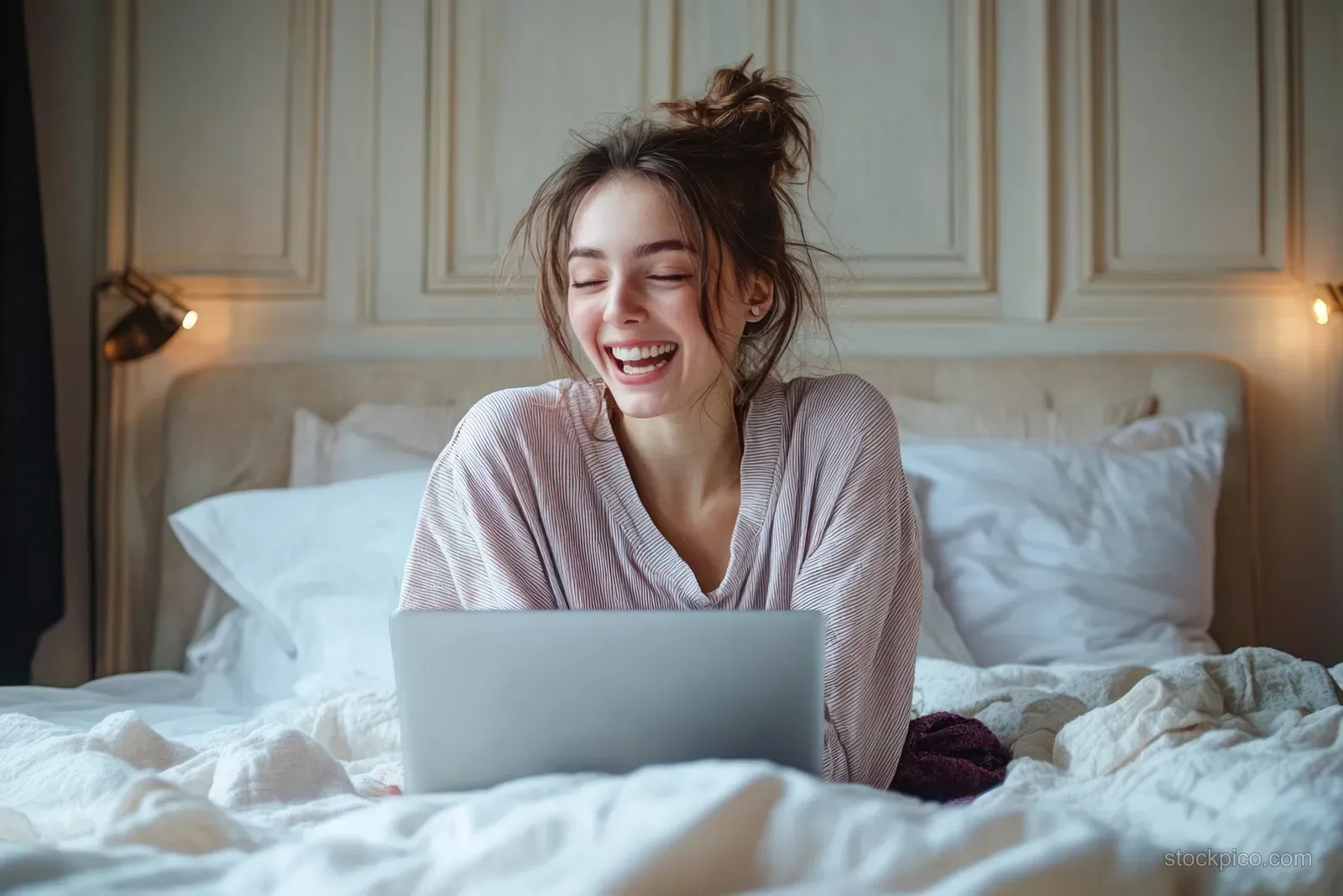 A smiling young woman uses a laptop while relaxing on a bed with soft pillows in warm bedroom light.