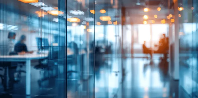 Blurred coworkers move behind glass partitions in a warmly lit corporate office with desks and meeting areas.