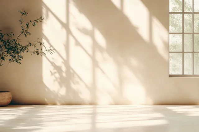 Sunlit studio corner with peach beige wall, window light casting leaf shadows onto a pale concrete floor.