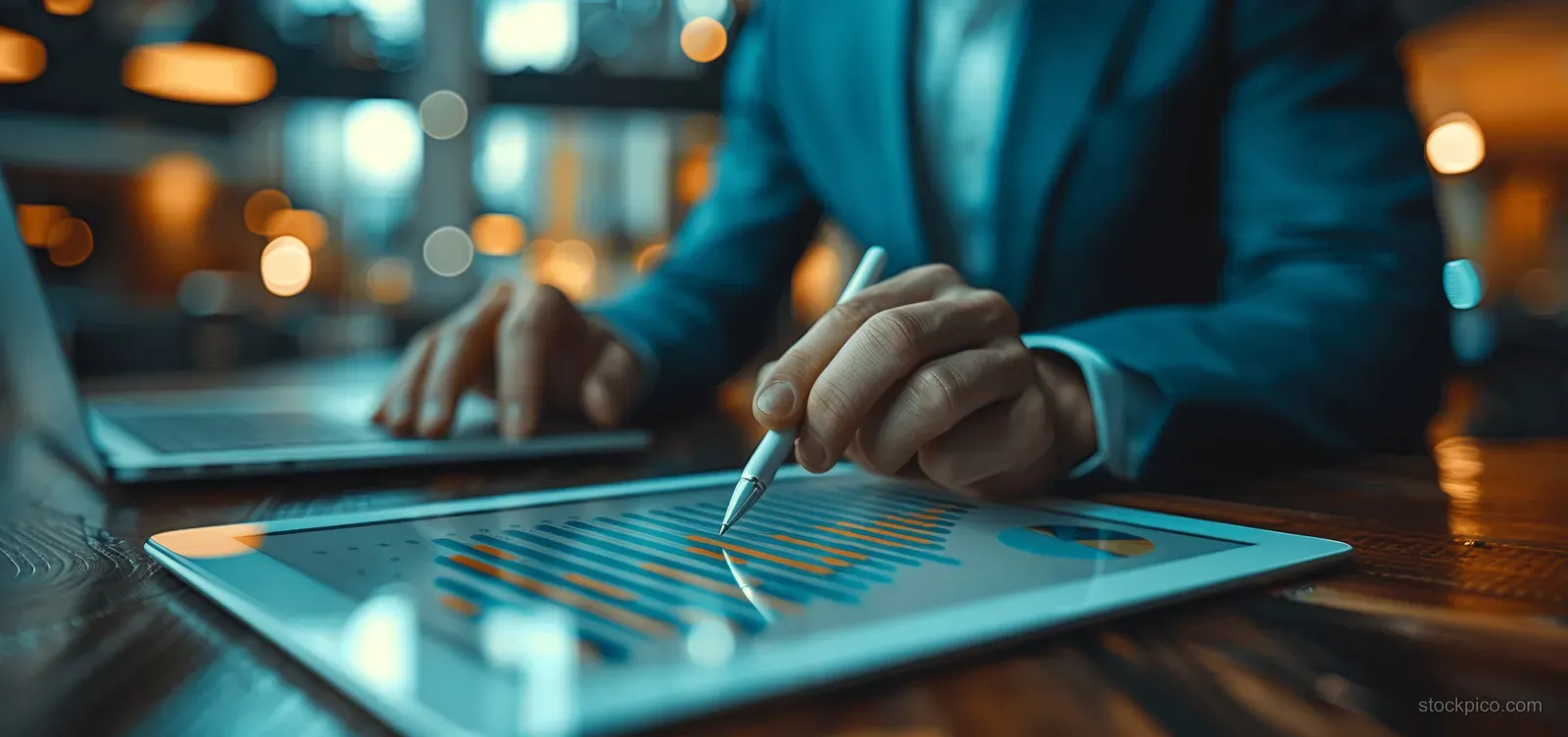 Two executives discuss project plans, pointing at a tablet chart while a laptop sits on an office desk.