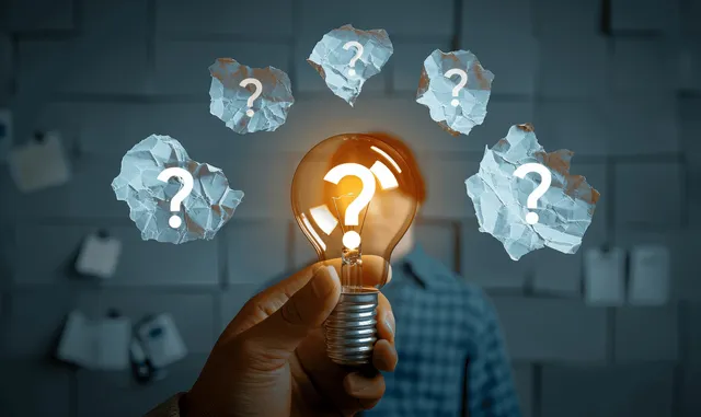 A hand holds a glowing lightbulb with torn paper pieces covered in question marks in front of a blurred office.