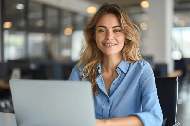 Smiling middle-aged businesswoman in blue shirt typing on laptop in modern office with Leadership And Management