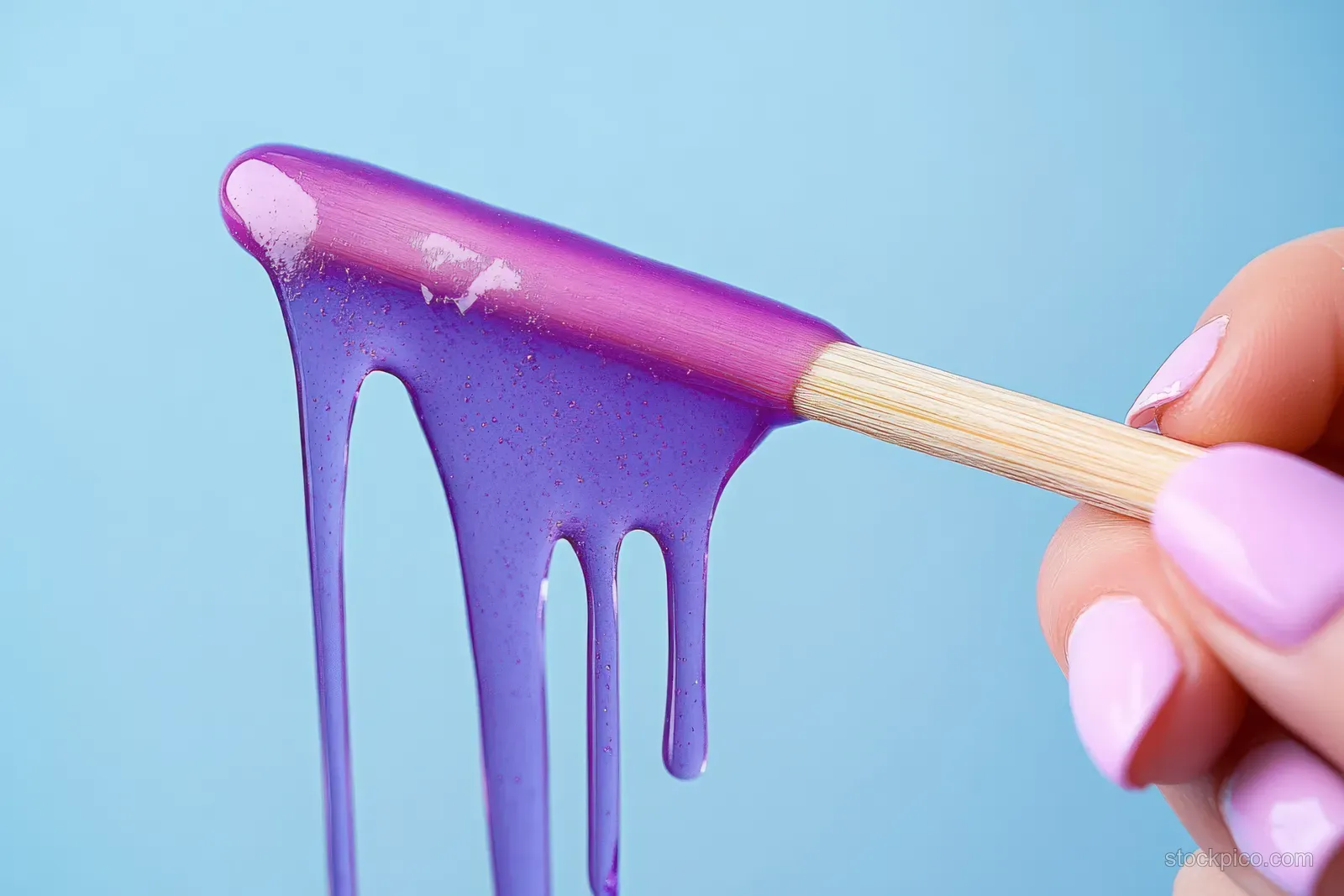 A hand uses a wooden spatula to spread glossy purple wax with visible drips on a blue background.
