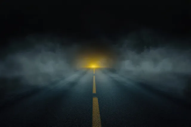 An empty dark road at night disappears into thick fog, with a fading yellow center line and distant hazy light.