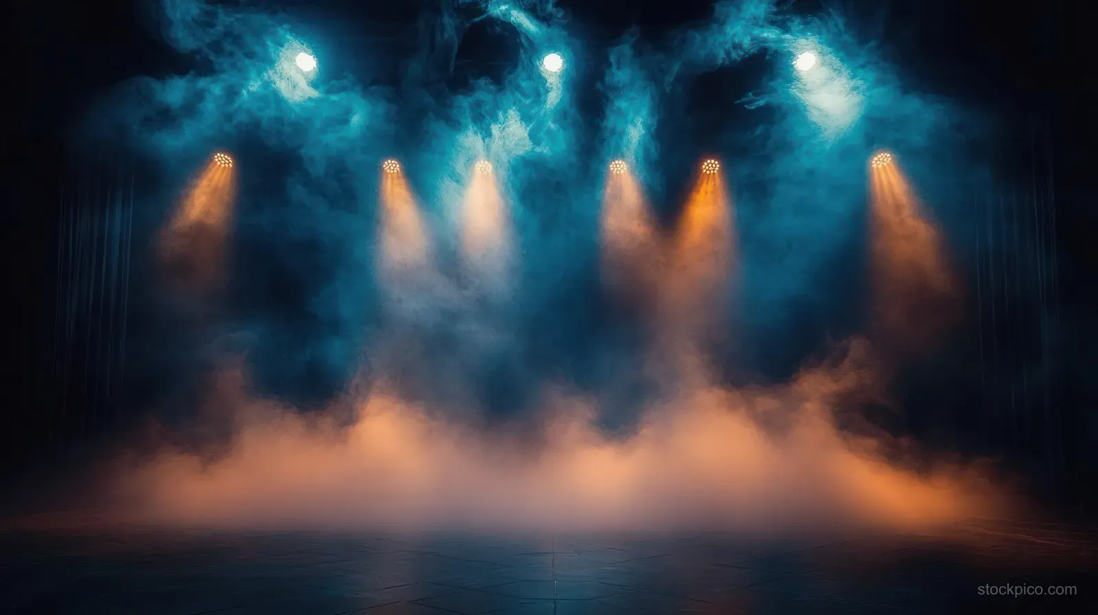 Blue spotlights shine through dense fog on a dark concert stage, forming glowing beams.
