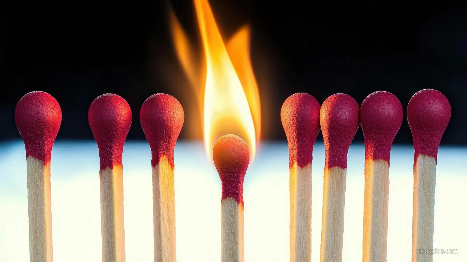 Nine aligned matchsticks with red tips, one burning with an orange flame between dark and light areas.