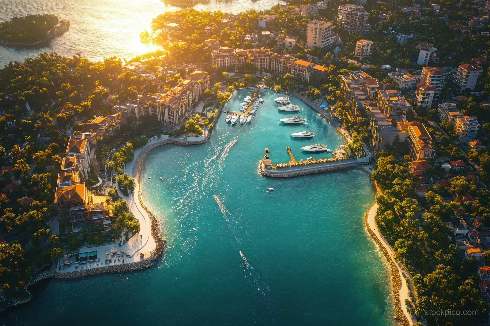 Aerial view of a sunlit coastal marina with docked yachts on turquoise water beside trees and waterfront buildings.