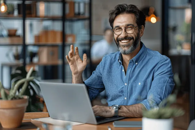 Smiling man in blue shirt video-calling from office desk, gesturing excitedly beside laptop