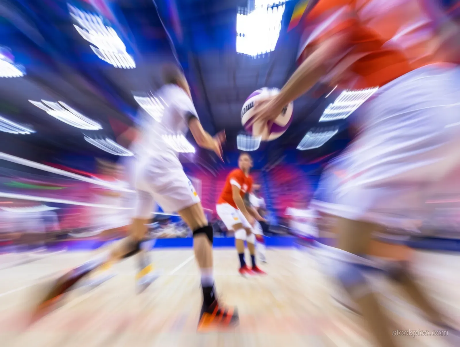 Men’s indoor volleyball players jump at the net for a block with visible motion blur in a bright arena.