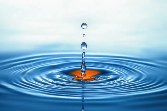 A clear water droplet falls into a blue pool, forming concentric ripples on the surface.