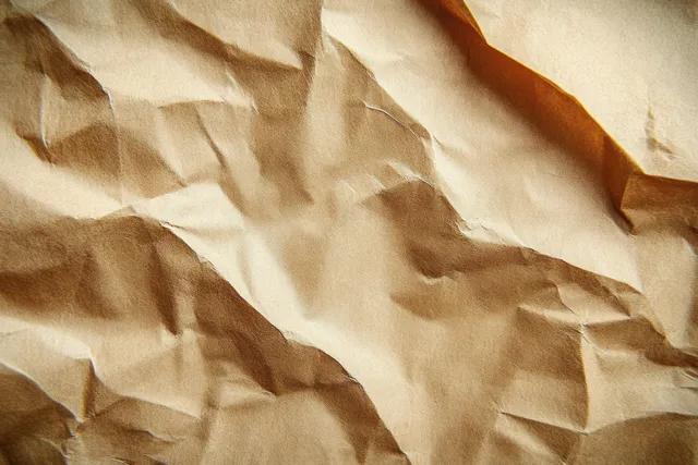 Crumpled brown kraft paper fills the frame with folded creases and soft shadows creating an abstract texture.